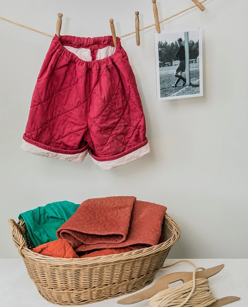 Selbstgenähte Fußballhose aus einer roten Steppdecke, daneben ein Foto mit jugendlichem Torwart in einer solchen Hose Anfang der 1950er Jahre